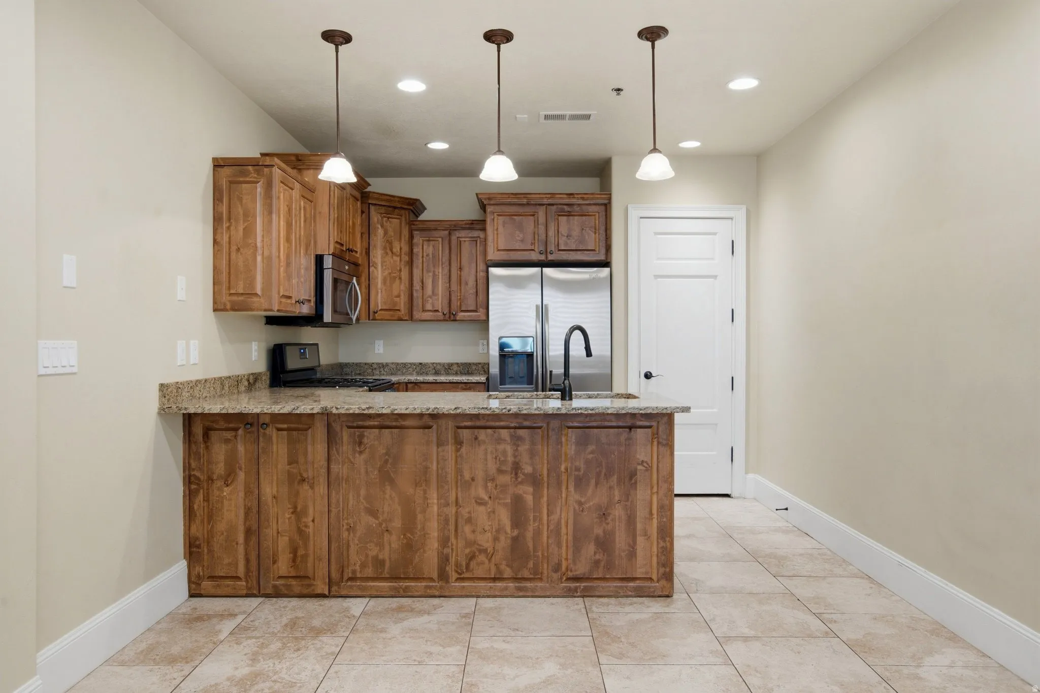 Kitchen featuring rich wood cabinetry, stone countertops, stainless steel appliances, a kitchen island with an undermount sink and a matte black faucet, and large format tile flooring