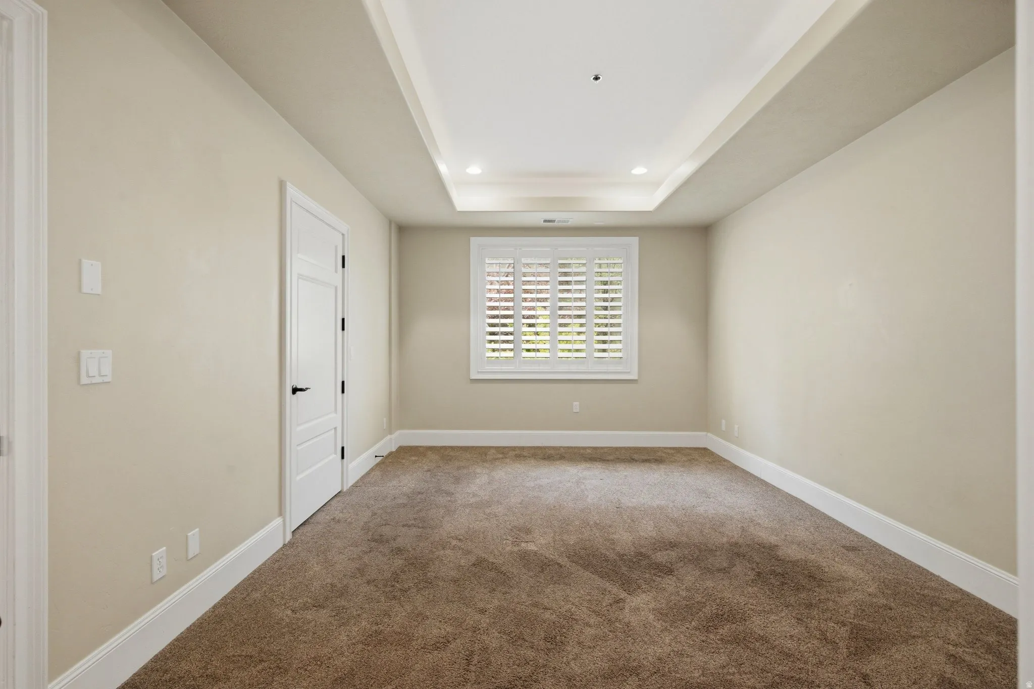 Recessed ceiling with integrated lighting, window with white plantation shutters, neutral wall tones, white trim, and brown carpeting