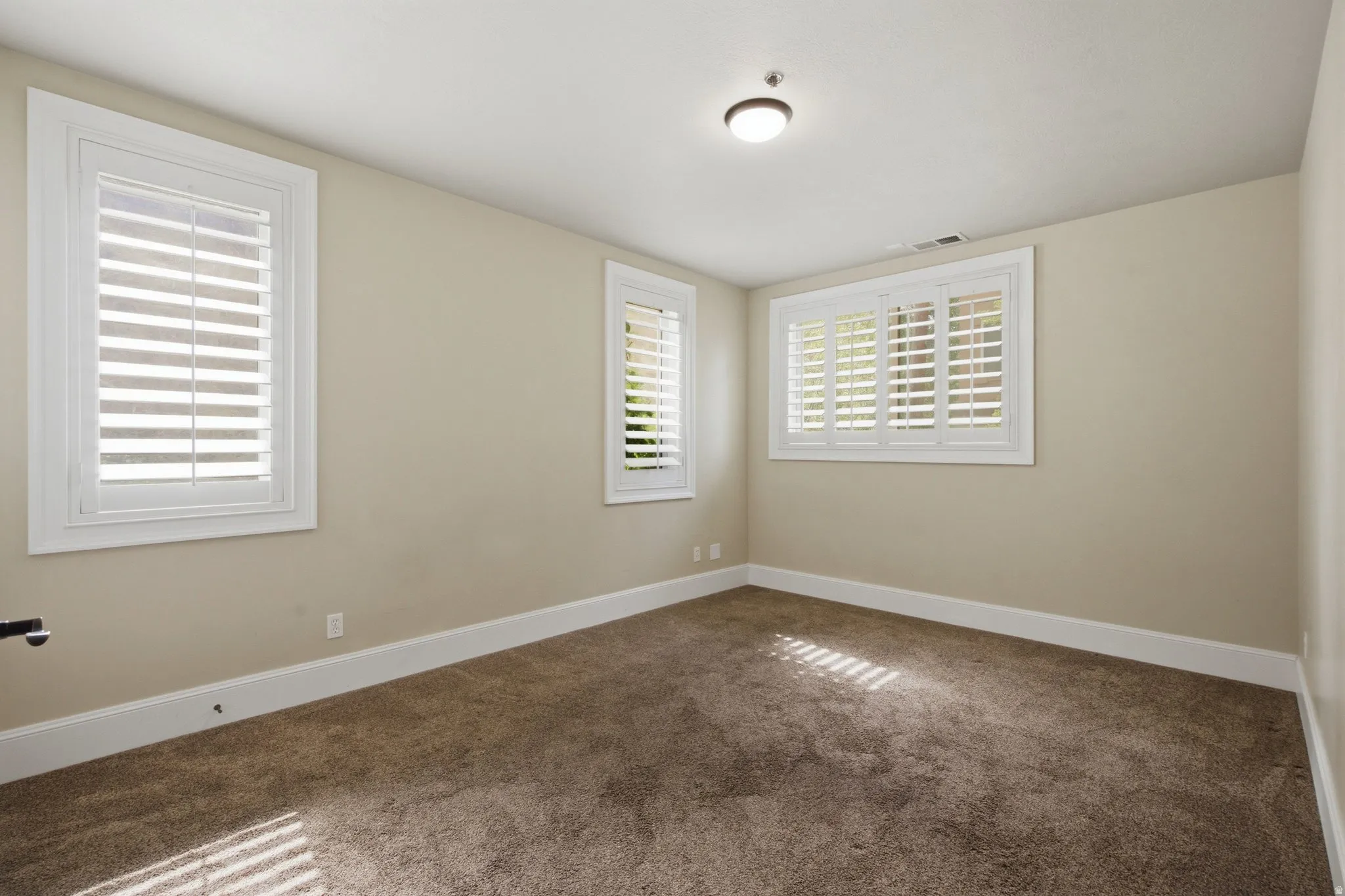 Room featuring three windows with white plantation shutters, neutral wall paint, and brown carpet flooring