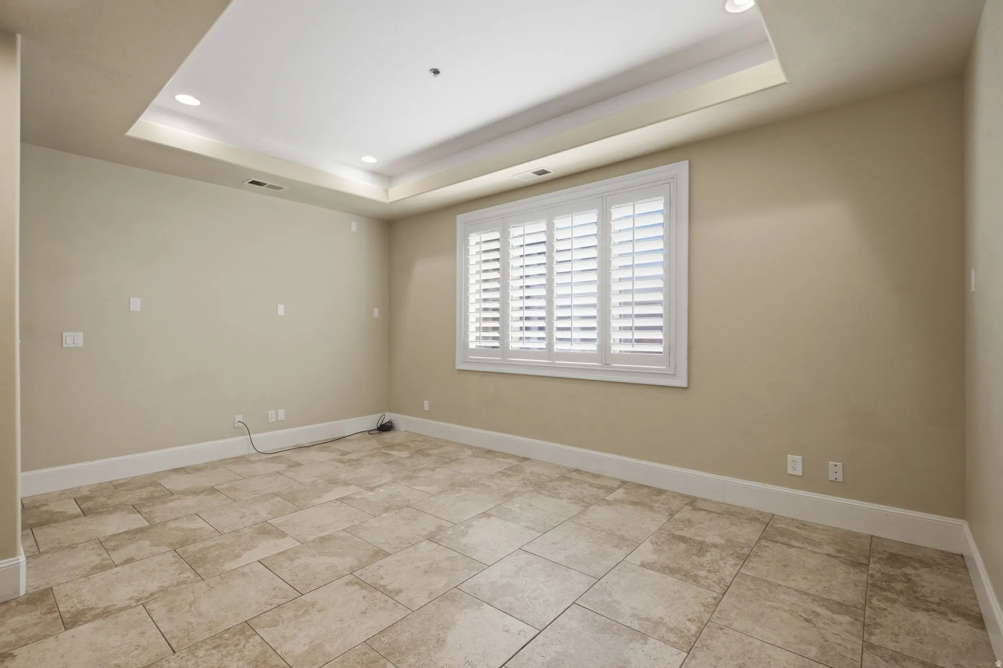 Spacious room featuring neutral wall tones, recessed lighting within a tray ceiling, and a window with white plantation shutters