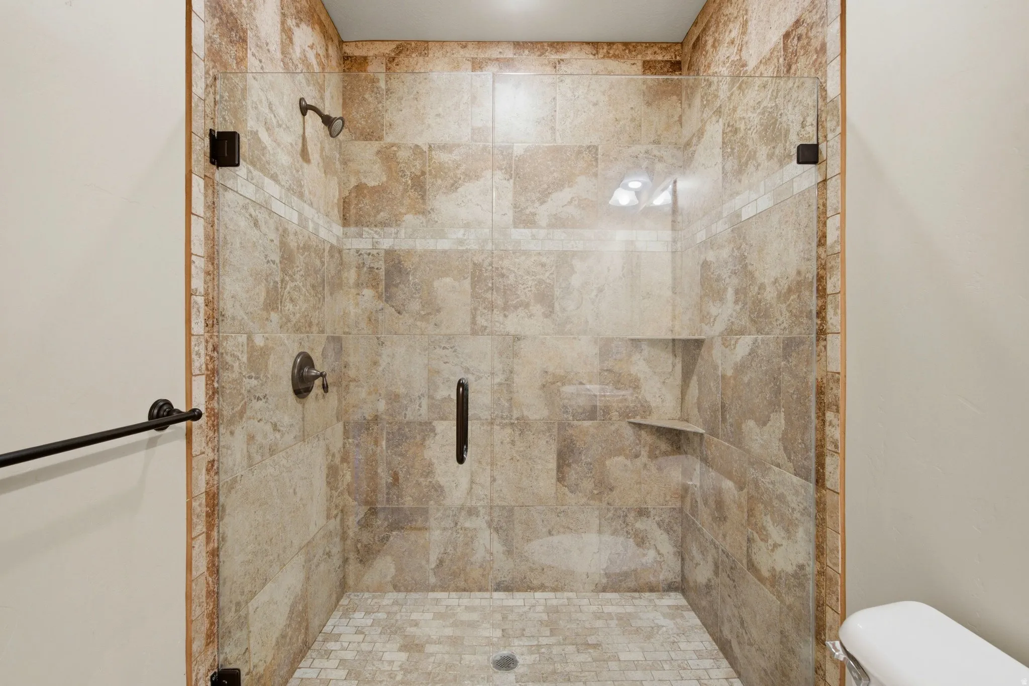 Walk-in shower featuring full-height natural stone-look tiling, a frameless glass enclosure, mosaic floor tiling, built-in shelving, and bronze-finish fixtures