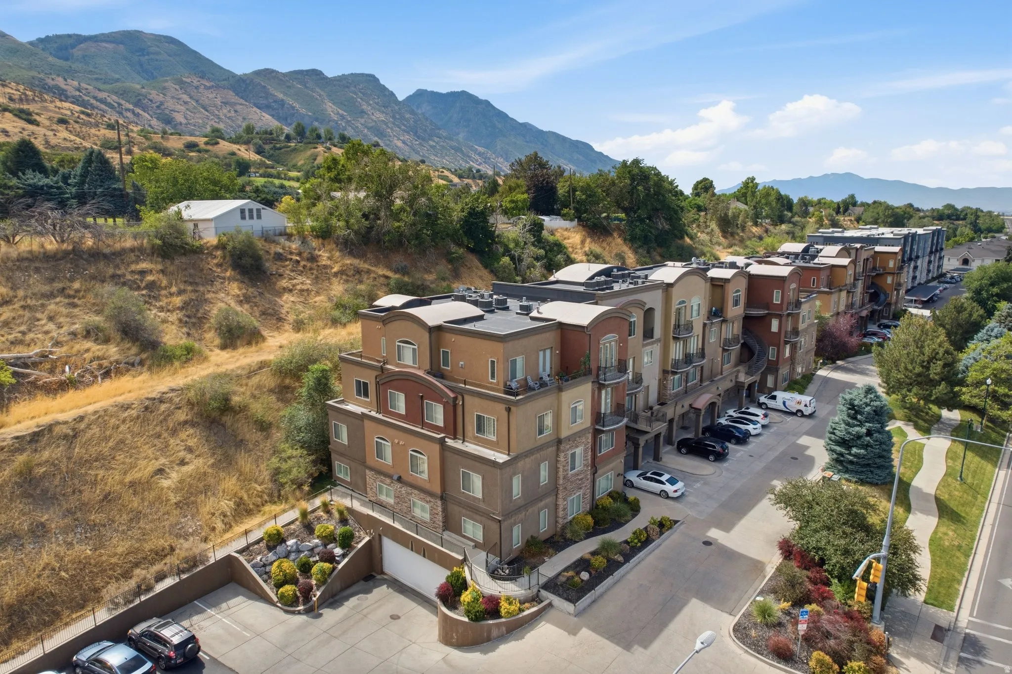 Multi-story residential building featuring balconies, a paved parking area, landscaping, and a mountainous backdrop