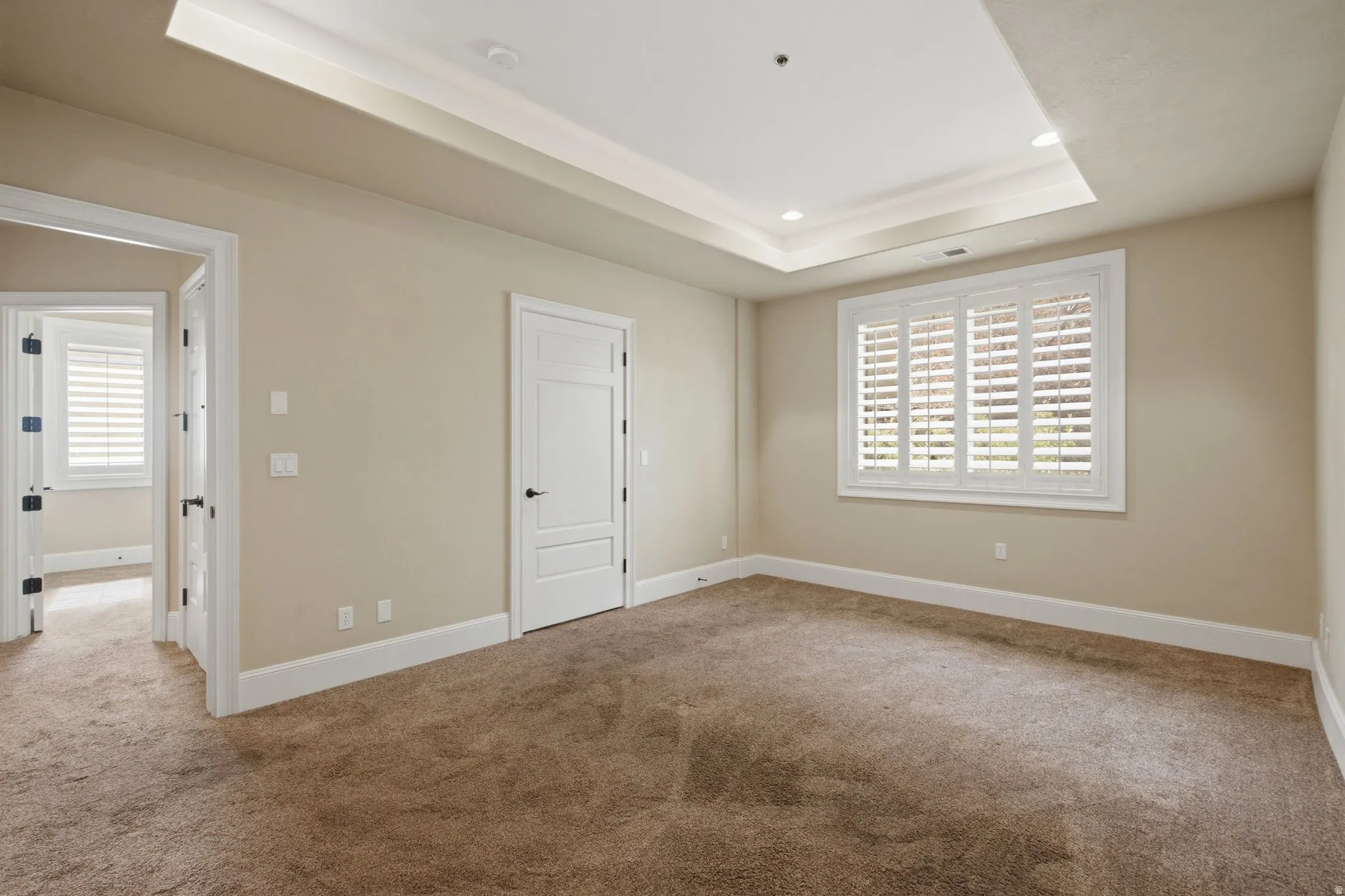 Spacious room featuring a tray ceiling with recessed lighting, plush carpet flooring, and a large window with plantation shutters