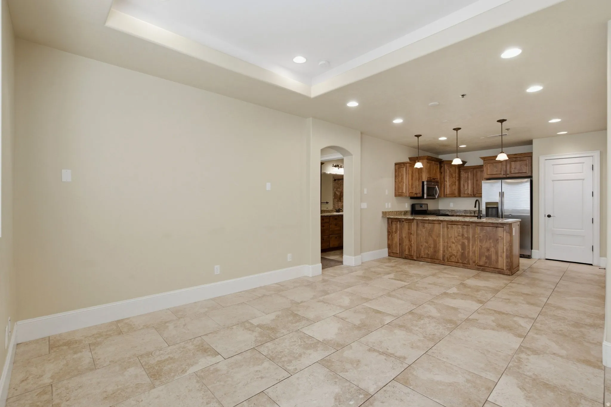 Open-concept living area featuring large format tile flooring, a tray ceiling with recessed lighting, and a kitchen with wood cabinetry, stainless steel appliances, and pendant lighting