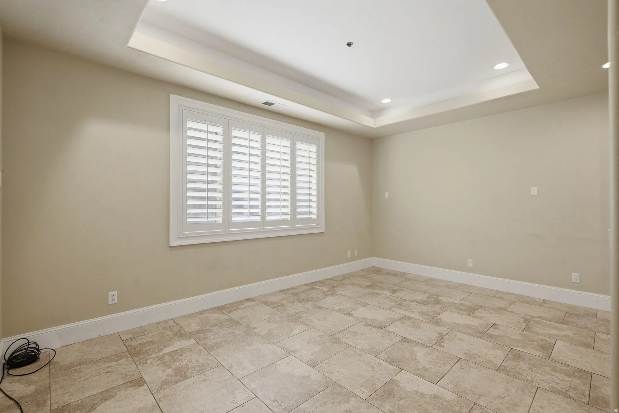 Spacious room featuring large format tile flooring, a tray ceiling with recessed lighting, and a window fitted with white plantation shutters