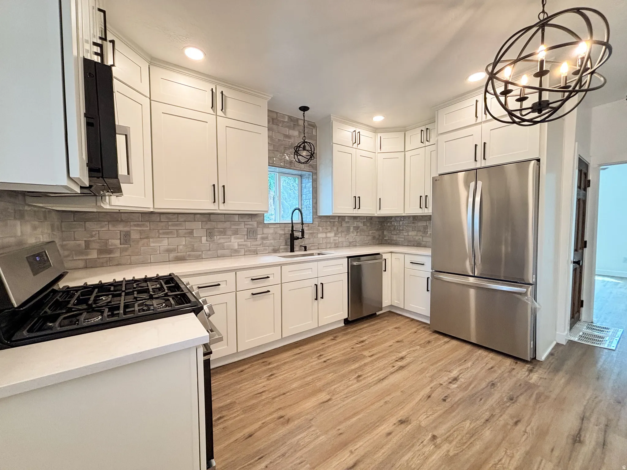 Modern kitchen featuring light wood-finish flooring, white shaker-style cabinetry with dark hardware, stainless steel appliances, a gray brick-patterned backsplash, and white countertops