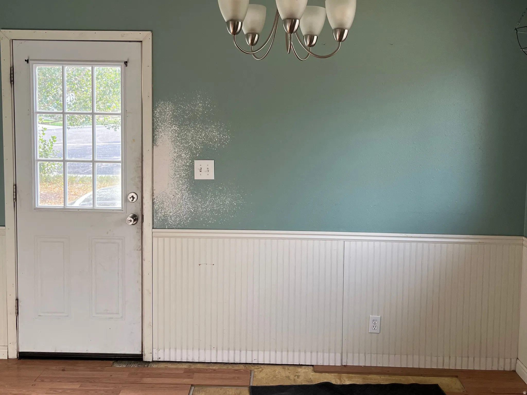 Glass-paneled door with a brushed nickel doorknob, white beadboard paneling, and a four-light chandelier. Floor needs fixed/replaced. This is the dining area and the side entrance from the concrete driveway.