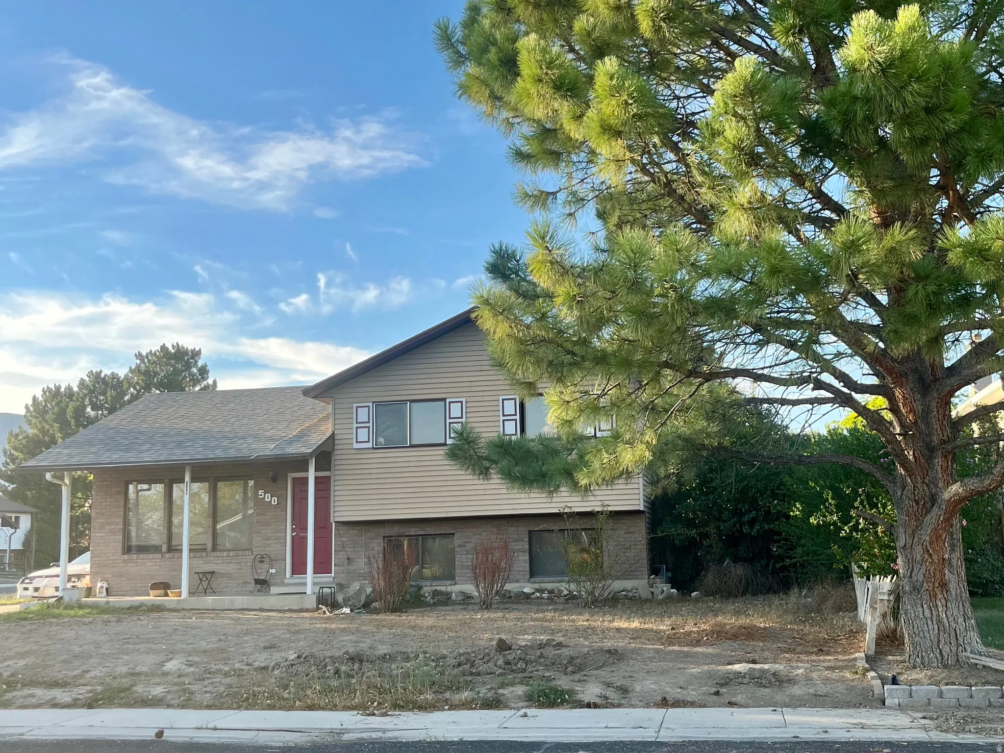 Split-level residence featuring brick and siding facade, a covered front porch, and a dark shingle roof. Large Pine Tree