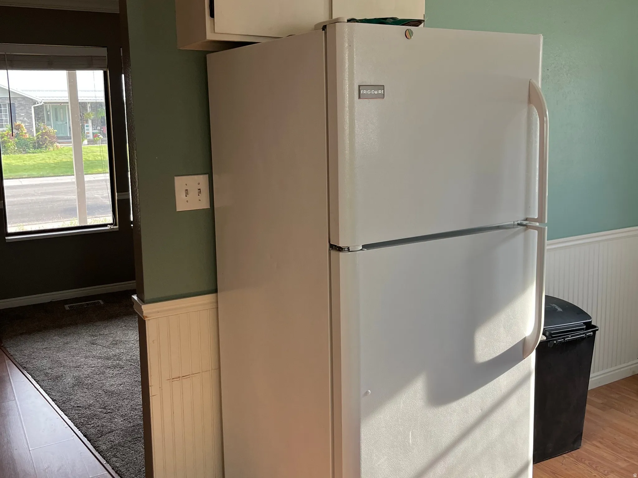 White top-freezer refrigerator, wood-finish flooring, beadboard wainscoting, and large window with blinds.