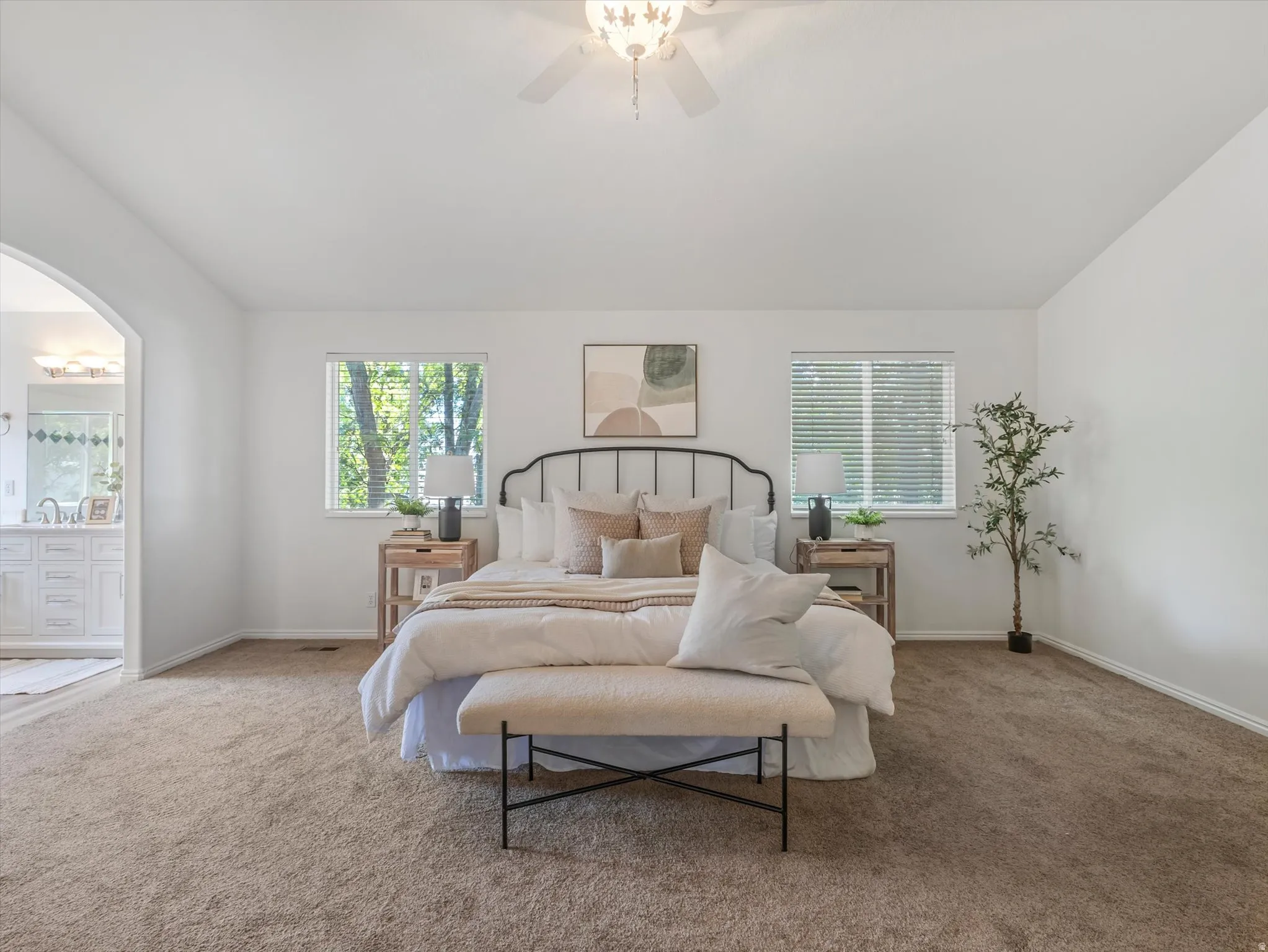 Carpeted bedroom featuring two windows with horizontal blinds, a ceiling fan with integrated lighting, and an arched doorway opening to a bathroom with a double vanity