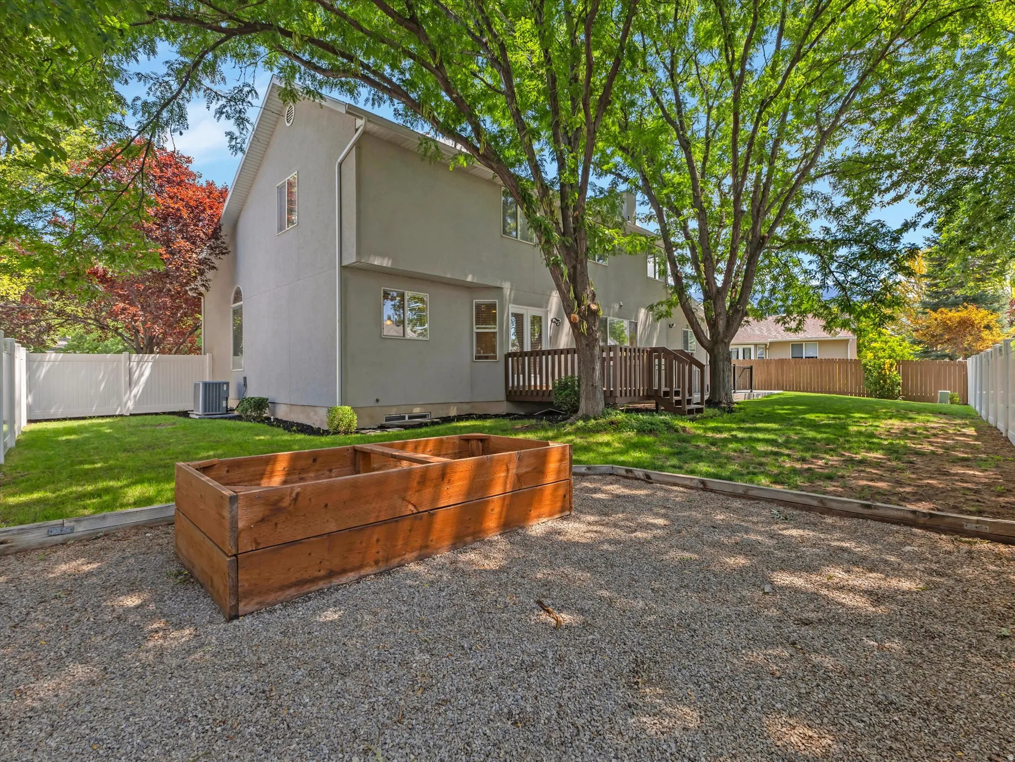 Spacious rear yard featuring a gravel area with a built-in planter box, a verdant lawn, a wood deck, a white privacy fence, and mature trees providing natural shade