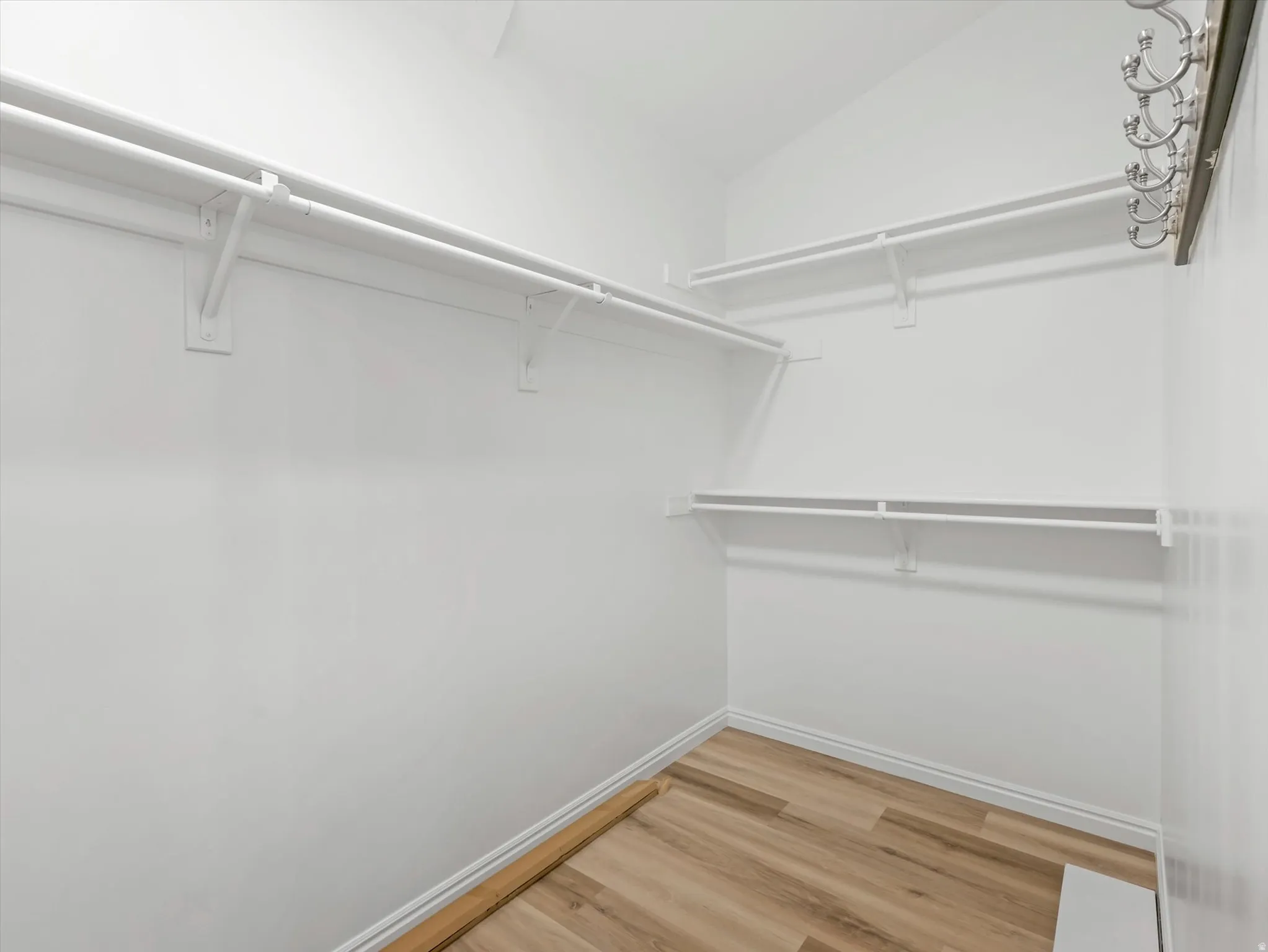 Primary walk-in closet featuring light wood-finish flooring, white walls, multiple built-in shelving units, and a wall-mounted coat rack