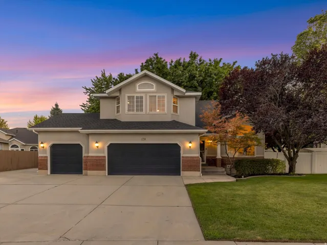 Two-story stucco and brick exterior featuring a three-car garage, mature trees, and a manicured lawn