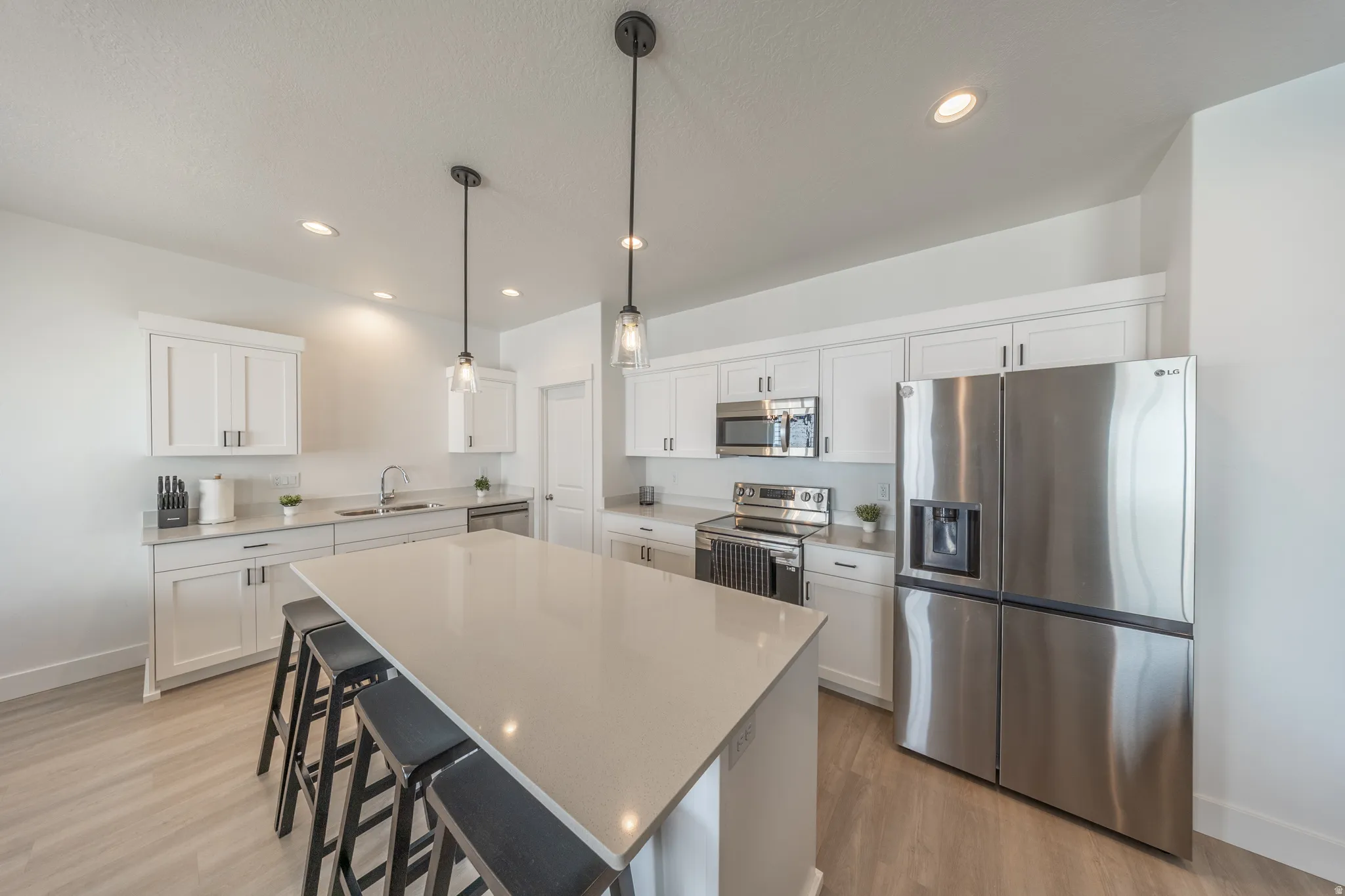 Bright kitchen featuring a substantial island with seating, white shaker cabinetry, stainless steel appliances, recessed lighting, and wood-finish flooring