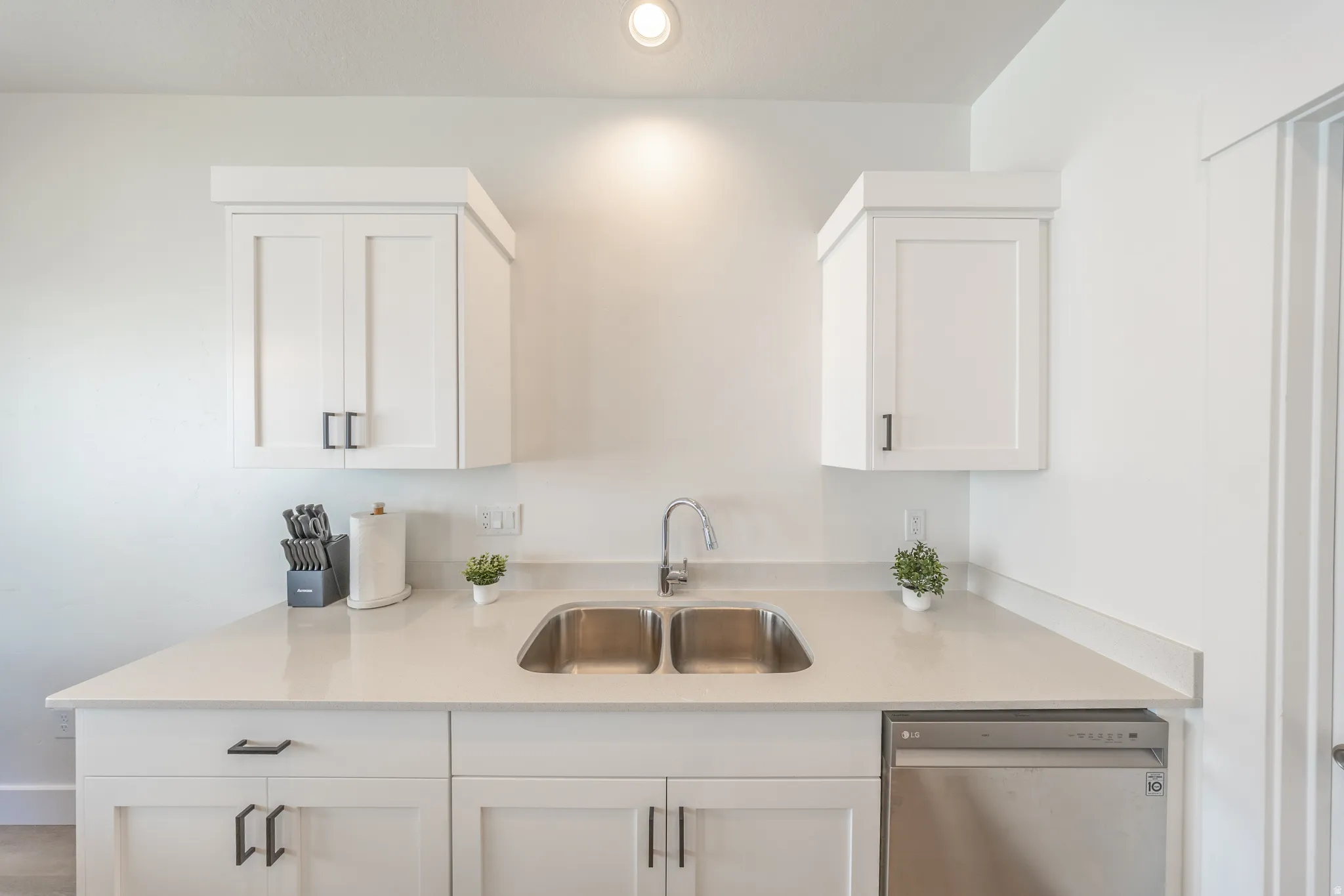 Bright kitchen featuring white shaker cabinetry, light-toned countertops, a double basin stainless steel sink with a gooseneck faucet, and a stainless steel dishwasher