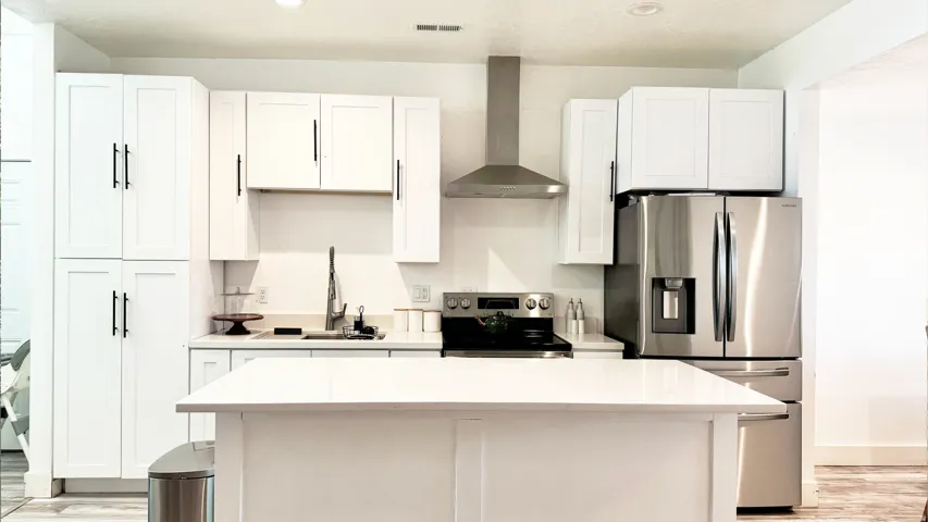 Contemporary kitchen featuring an expansive center island with white countertop, white shaker cabinetry with matte black hardware, stainless steel appliances, and a stainless steel range hood