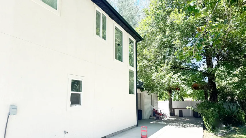 Exterior featuring white stucco siding, contrasting dark trim, multiple large windows, and a shaded concrete patio area