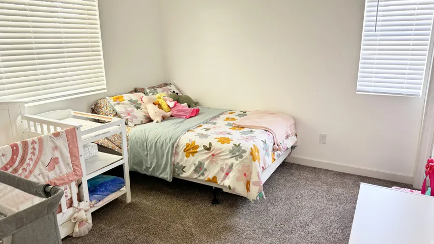 Bedroom featuring light grey carpet flooring, white walls, and two windows with horizontal blinds