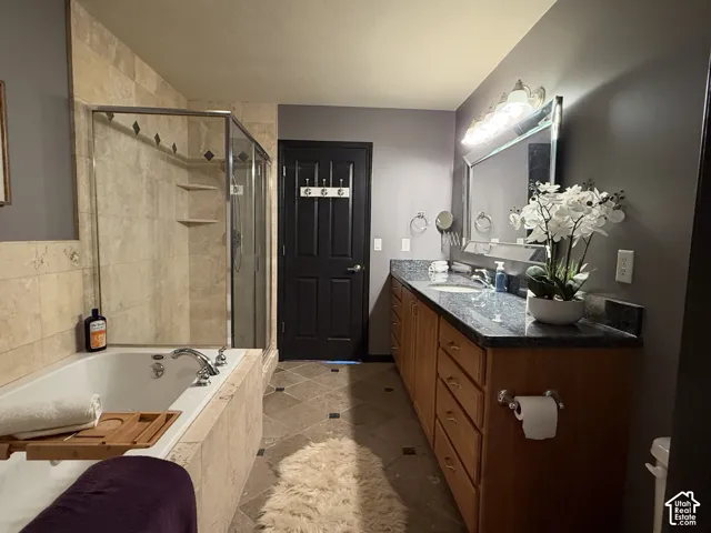 Bathroom featuring a dual-sink vanity with dark stone countertops, natural stone tile flooring, a soaking tub with stone tile surround, and a glass-enclosed shower with tiled walls