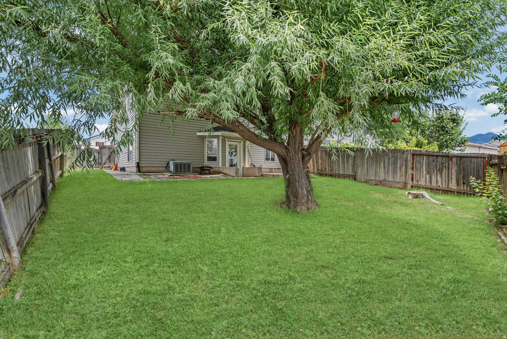 Expansive fenced backyard featuring a mature deciduous tree, a manicured lawn, a concrete patio, and a visible air conditioning unit