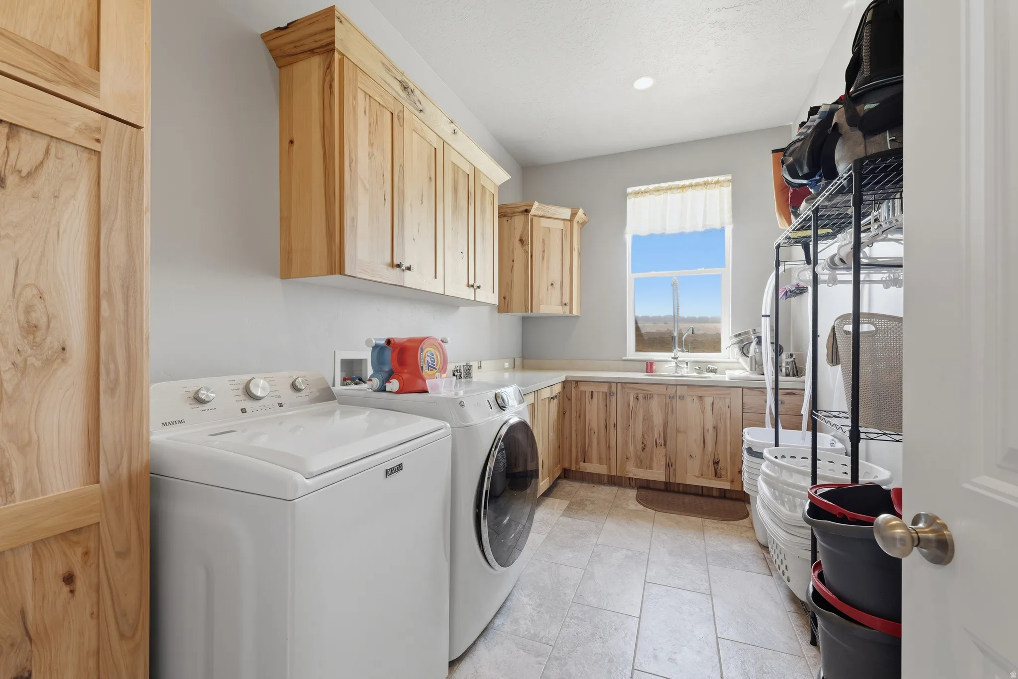 Laundry room featuring natural wood cabinetry, light grey wall paint, recessed lighting, a window with a sink beneath, and tile flooring