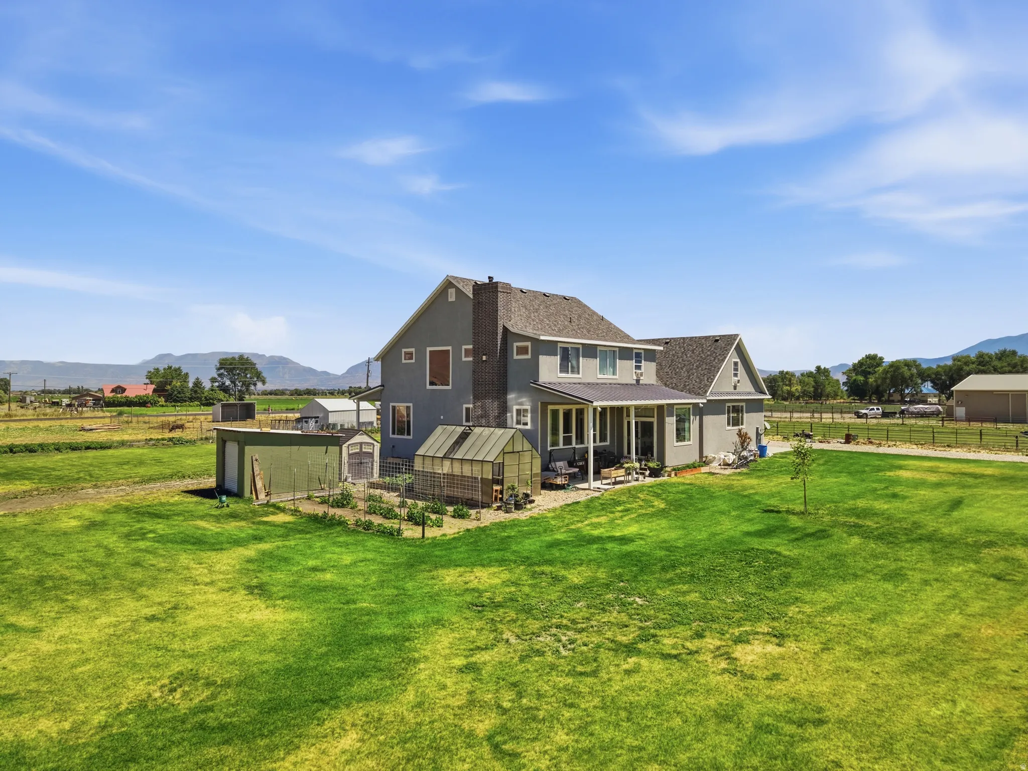 Expansive property featuring a grey two-story residence with a covered patio, adjacent greenhouse, and multiple outbuildings