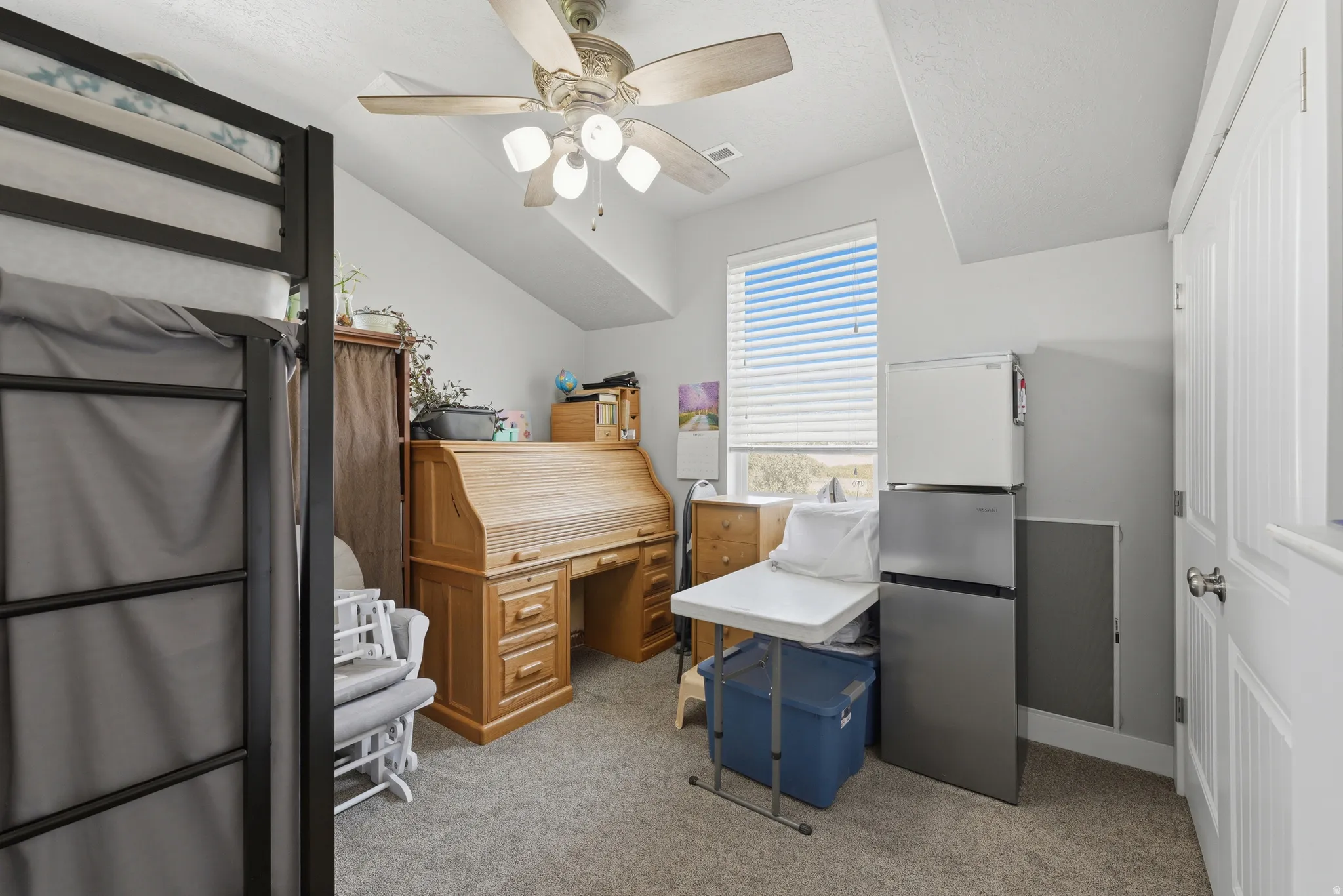 Carpeted interior space featuring a window with horizontal blinds, a ceiling fan with integrated lighting, and a white paneled door with a brushed nickel knob