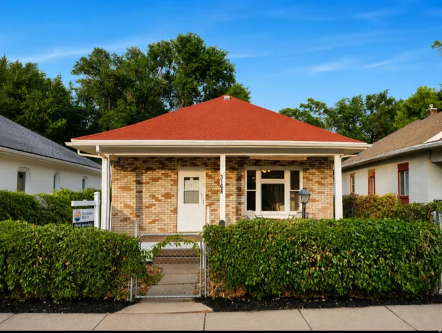 Brick facade home featuring a red asphalt shingle roof, covered front porch, white-framed windows, and a chain-link gate with established perimeter hedges