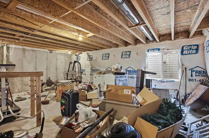 Unfinished basement featuring exposed wood joists, concrete foundation walls, and a window well with a louvered cover