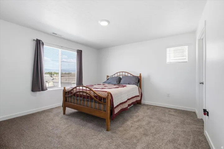 Bedroom featuring soft gray carpet flooring, a large window with blinds, a smaller window with blinds, white baseboards, and a white interior door