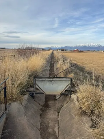 Concrete irrigation ditch with a metal water gate, bordered by dry grasses and fields, set against a distant mountain range