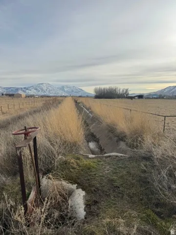 Expansive agricultural land featuring a visible irrigation ditch, a water control valve, mature dry grasses, and distant snow-capped mountains under a clear sky