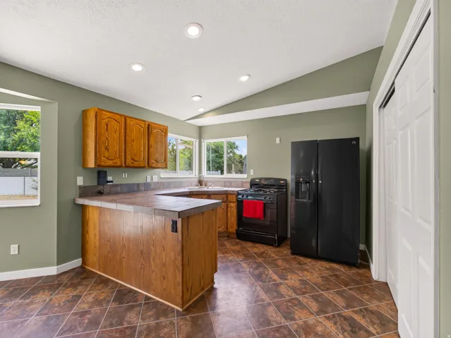 Kitchen featuring wood-finish cabinetry, tile-finish countertops, black appliances, recessed lighting, and a vaulted ceiling