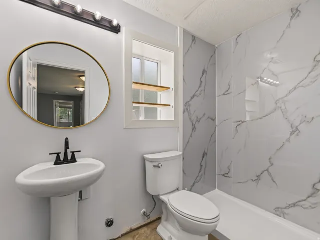 Bathroom featuring a pedestal sink with matte black fixtures, a gold-framed circular mirror, and a window with wood shelving