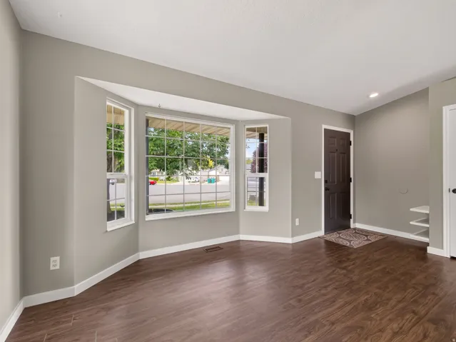 Spacious living area featuring a large bay window with grid patterns, wood-finish flooring, light grey wall paint, white baseboards, and a dark paneled entry door