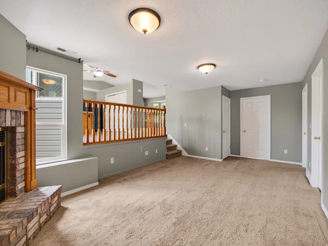 Spacious interior room featuring a brick fireplace with a wood mantel, light-colored carpeting, neutral wall finishes, and a built-in wood railing system