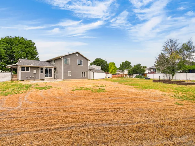 Expansive backyard featuring a large open area, a gray-sided residence with multiple windows, a covered patio, and raised garden beds