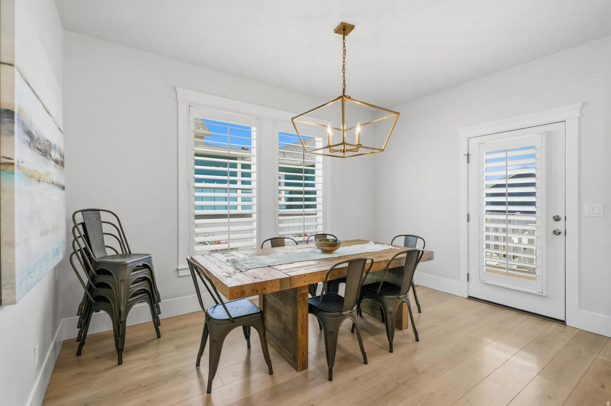 Dining area featuring wood-finish flooring, two large windows with plantation shutters, a glass-paneled door with plantation shutters, a brass-tone geometric chandelier, and white baseboards