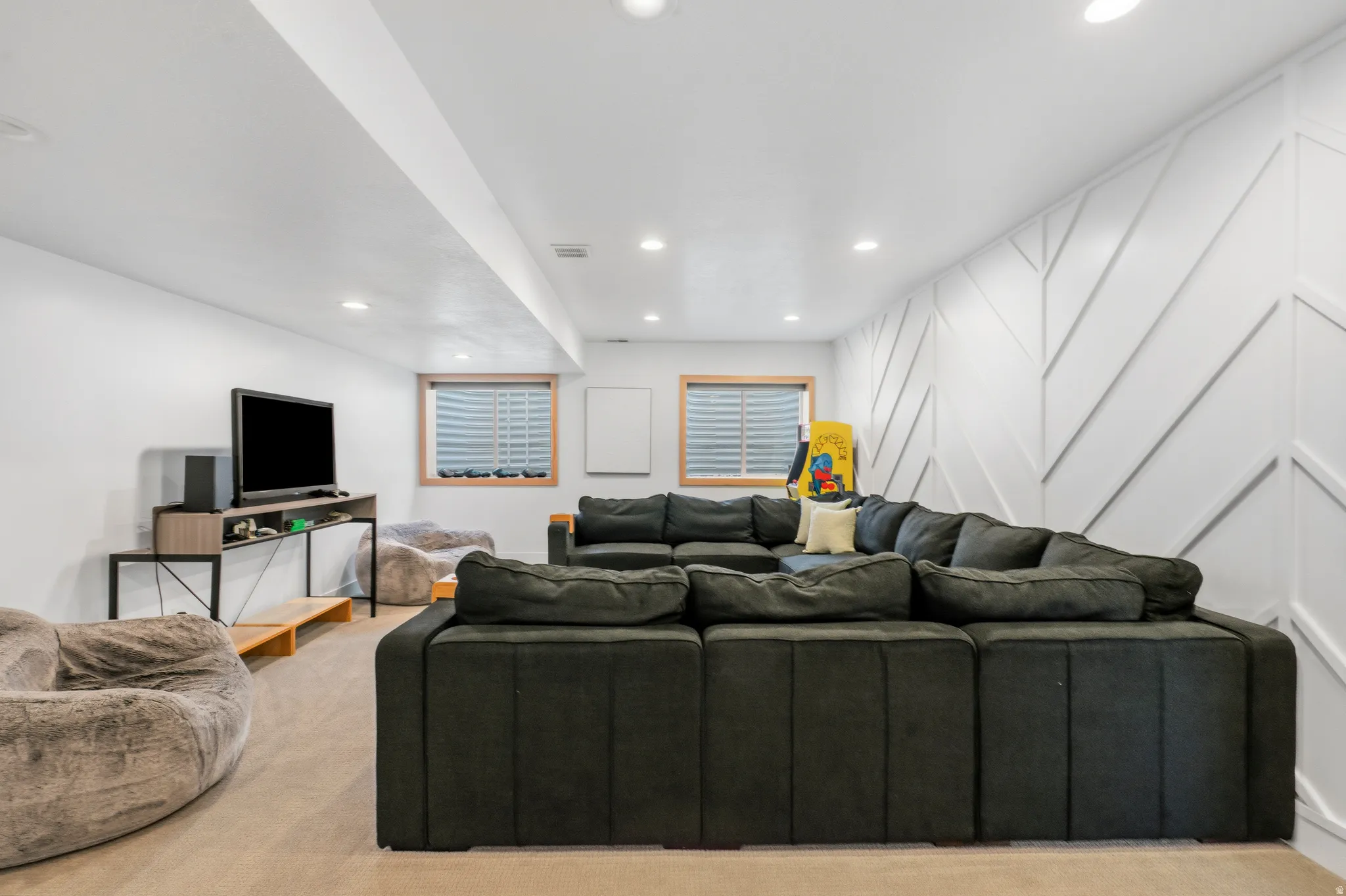 Finished basement lounge area featuring a decorative accent wall with geometric paneling, recessed lighting, and two windows with horizontal blinds