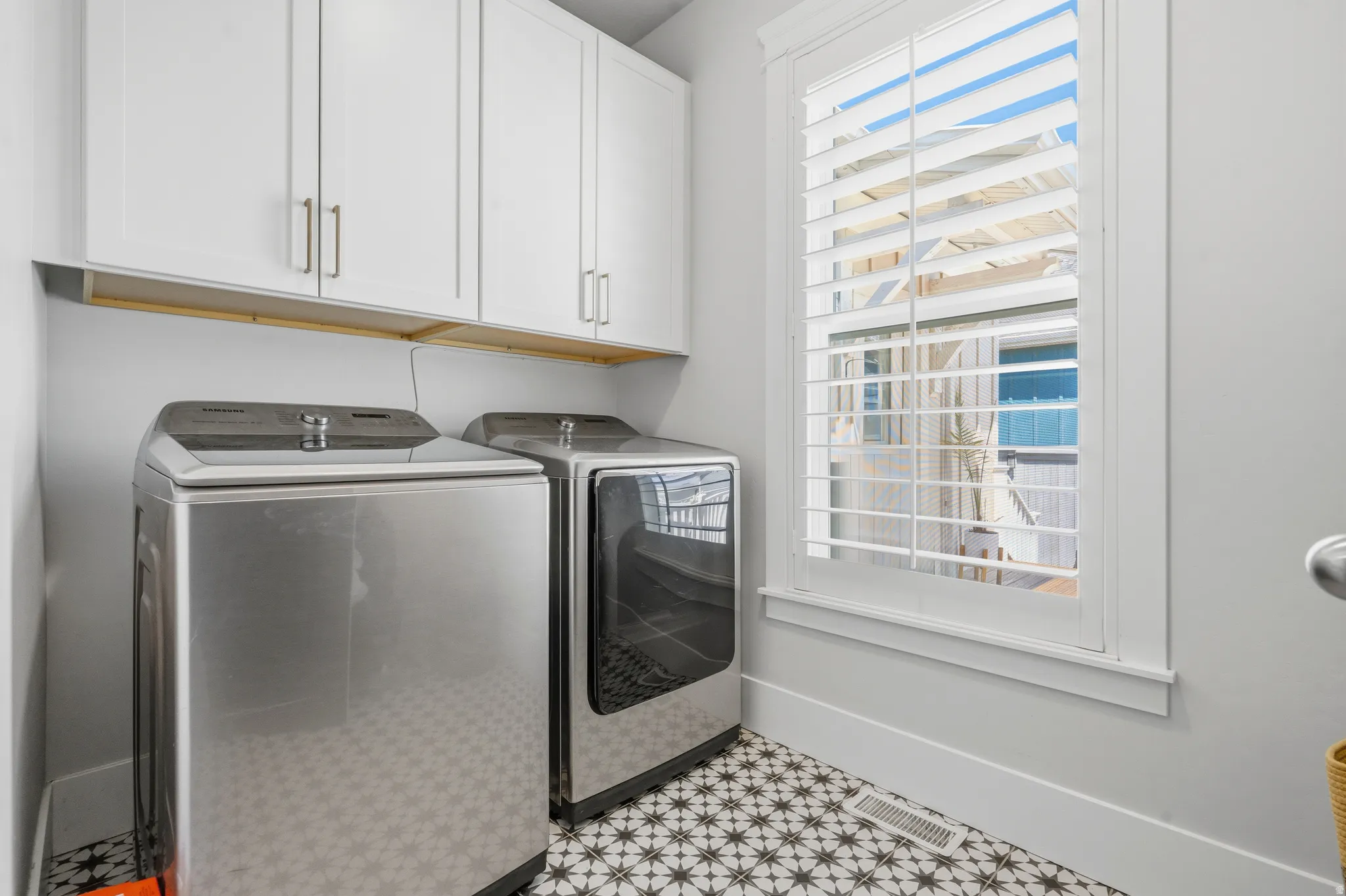 Laundry area featuring a window with white louvered shutters, patterned floor tiles, white shaker-style cabinetry with brushed nickel hardware, and an integrated wood shelf