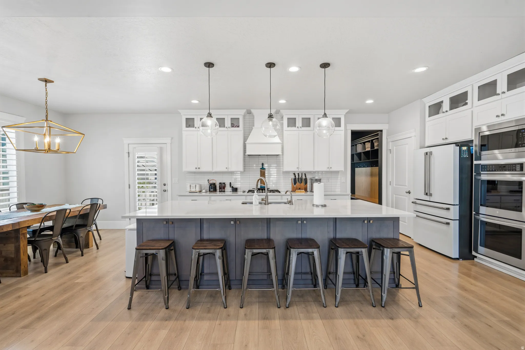 Spacious kitchen featuring a large central island with seating, white shaker cabinetry, stainless steel appliances, pendant lighting, and wood-finish flooring