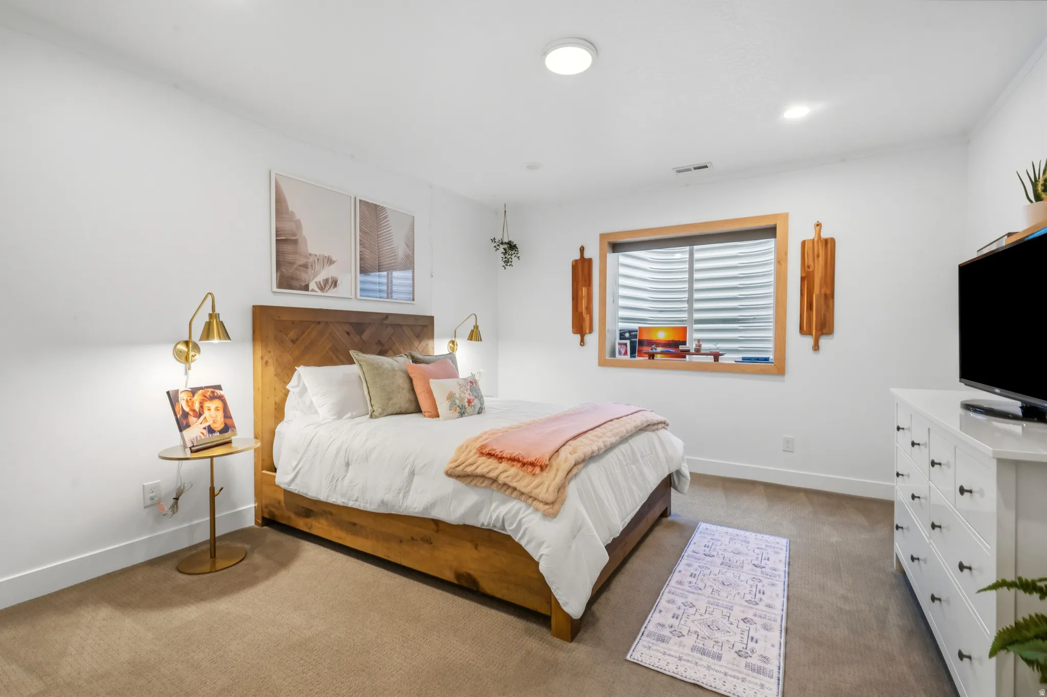 Carpeted room featuring white walls, a wood-finish bed frame with a herringbone headboard, two brass-finish wall sconces, and a window with wood trim