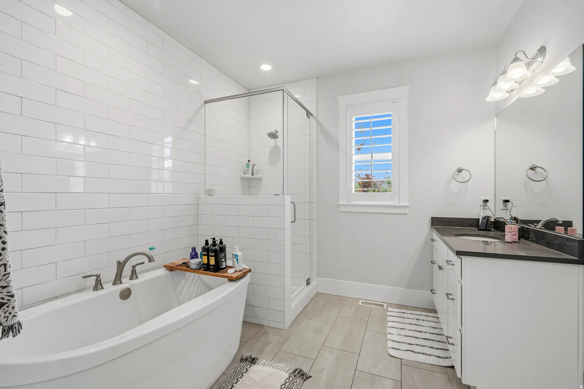 Freestanding soaking tub with a wall of white subway tile, an enclosed glass shower, a vanity with a dark countertop, and light-toned tile flooring
