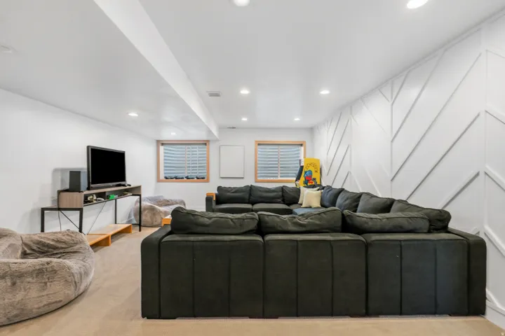 Finished basement lounge area featuring a decorative accent wall with geometric paneling, recessed lighting, and two windows with horizontal blinds