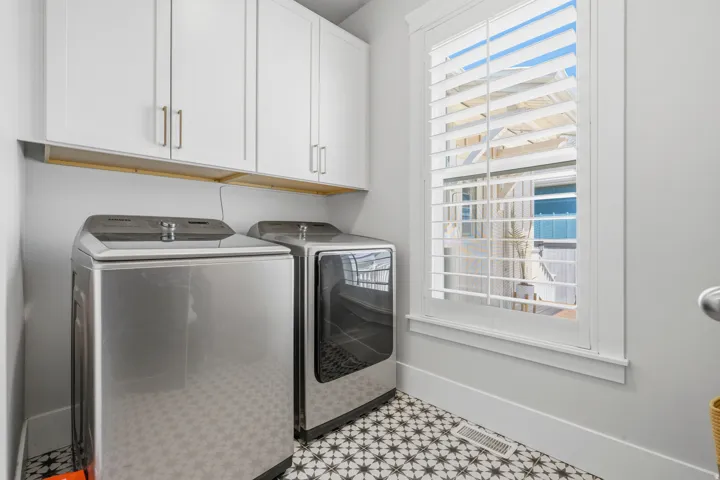 Laundry area featuring a window with white louvered shutters, patterned floor tiles, white shaker-style cabinetry with brushed nickel hardware, and an integrated wood shelf
