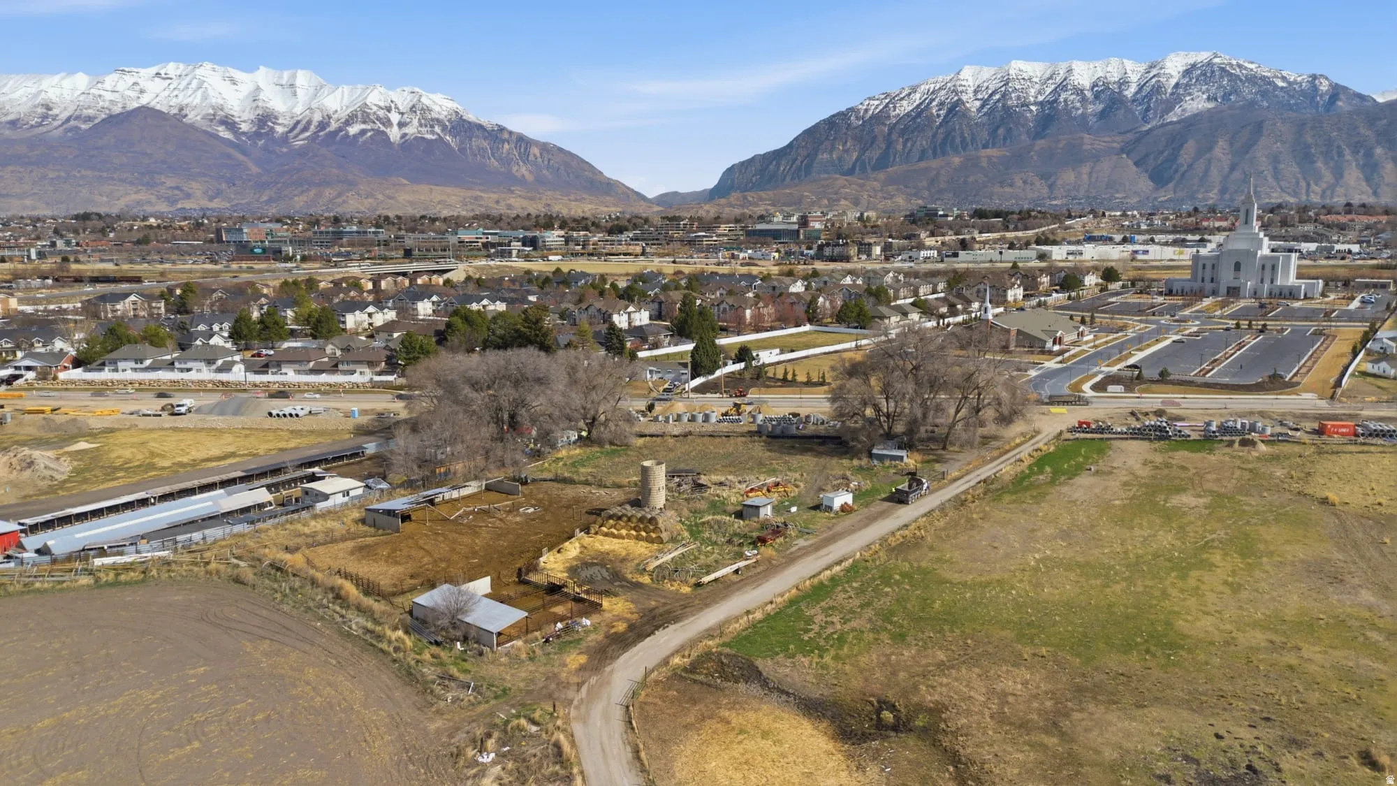 Expansive undeveloped land featuring a dirt road, multiple outbuildings, and a silo, framed by a snow-capped mountain range and distant urban development