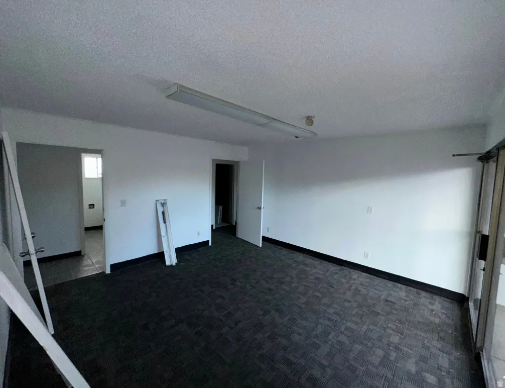 Spacious room featuring dark patterned carpeting, white painted walls, and a dropped ceiling with fluorescent lighting