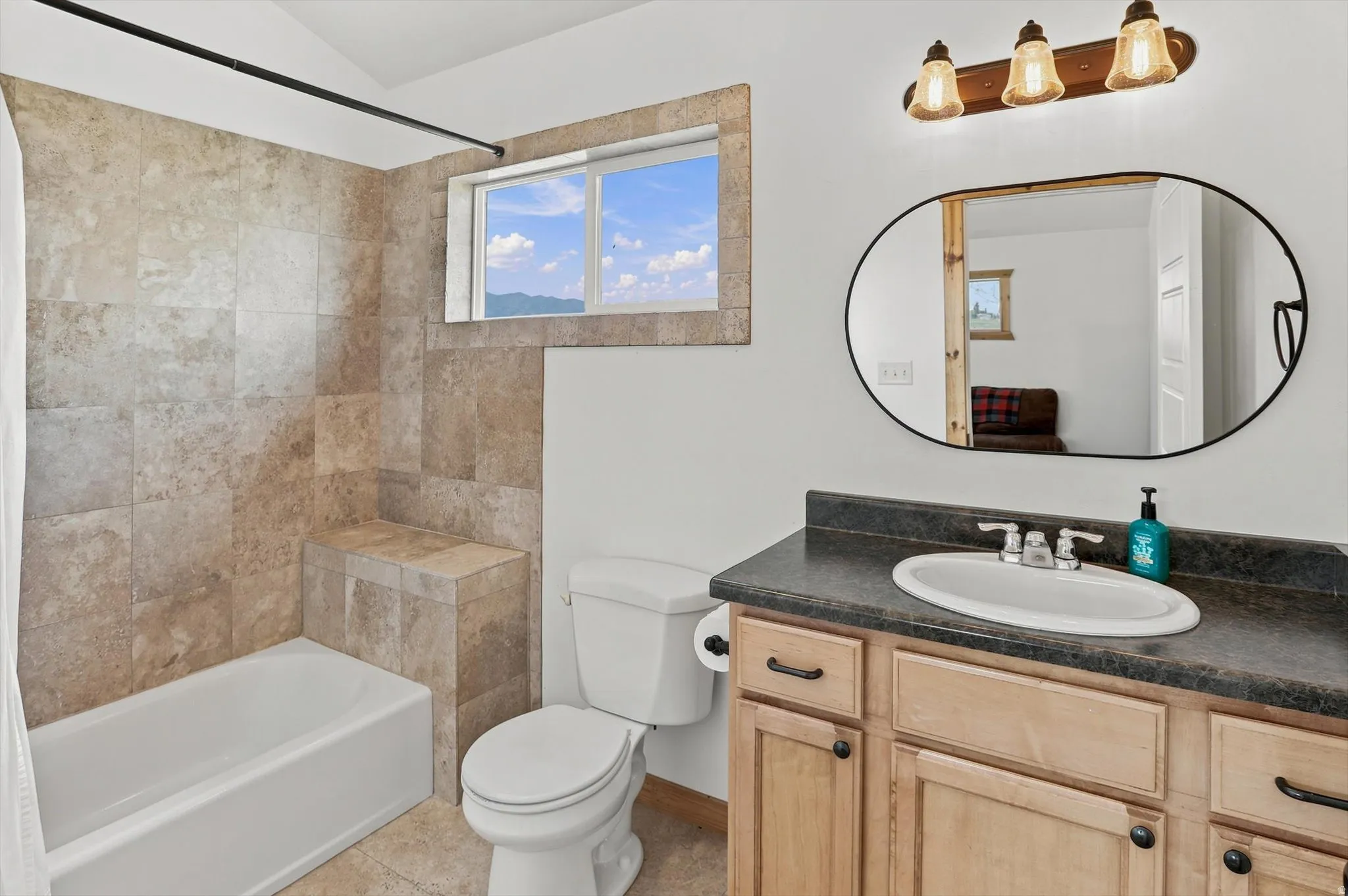 Bathroom featuring a tiled tub and shower enclosure with a built-in bench, a window, a single vanity with a dark countertop, an oval mirror, and a three-light fixture