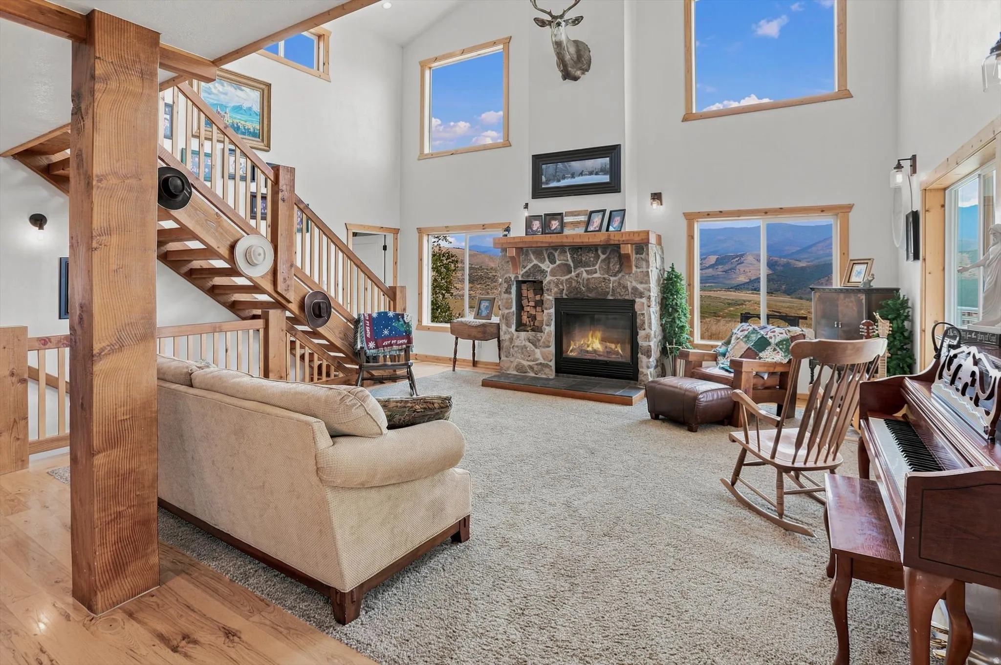 Open-concept living area featuring a stone fireplace with a wood mantle, high ceilings, multiple windows, and a wood-finish staircase