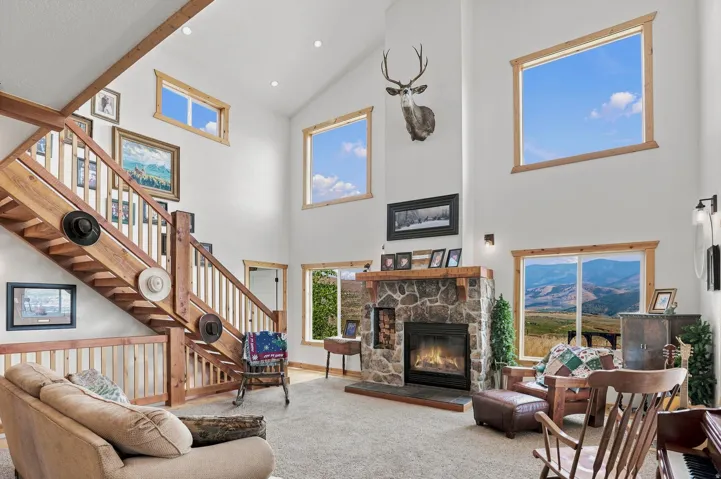 Vaulted living space featuring a stone-clad fireplace with mantel, an open-tread wood staircase, and multiple windows with wood trim